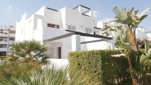 Un edificio moderno blanco con balcones, rodeado de vegetación, incluyendo palmeras y un seto.