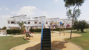 Un parque infantil con un tobogán azul y columpios, rodeado de césped cuidado y un edificio con varios balcones al fondo.