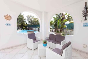 Un patio techado con mobiliario de mimbre blanco, una mesita con una planta y vistas a una piscina y follaje.