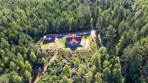 Vue aérienne d’une propriété entourée d’une forêt dense. On y voit deux bâtiments principaux avec des toits rouges, une allée et un petit espace de stationnement.