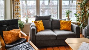 Un salon chaleureux avec un canapé gris, un fauteuil en cuir noir et une table en bois. Deux coussins jaunes sont posés sur le canapé, et une plante en pot se trouve sur le rebord de la fenêtre.