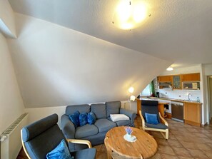 Una sala de estar acogedora con un sofá, dos sillones y una mesa de centro de madera. Se ve una cocina al fondo, con armarios de madera y un microondas.