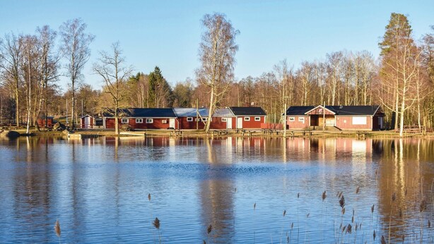 A row of red wooden cabins by a calm lake with reflections, surrounded by trees.