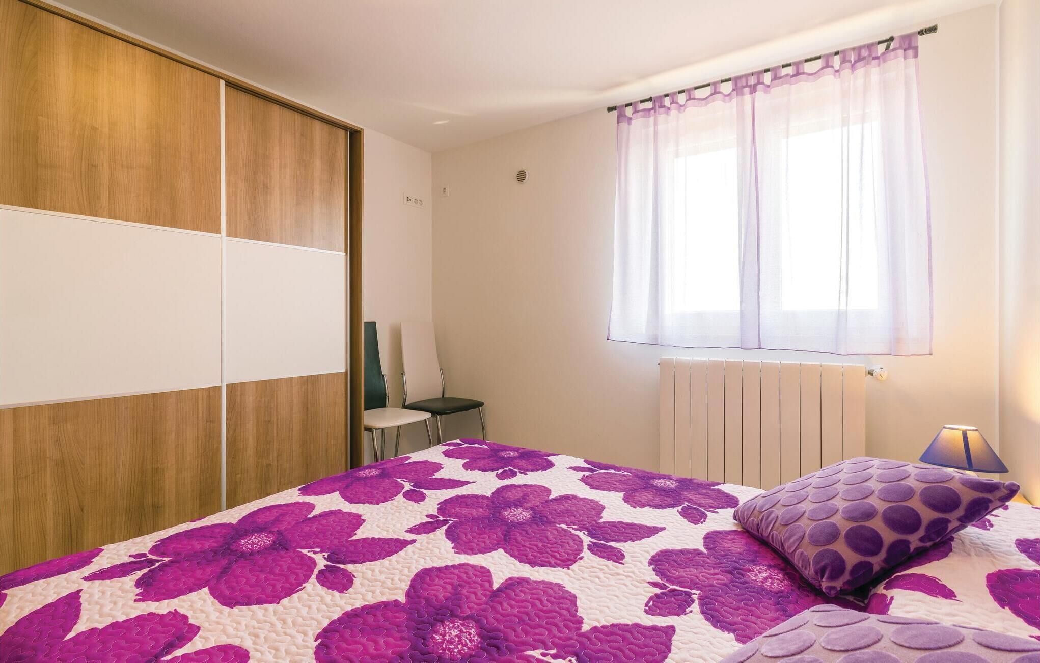 A bedroom with a bed, a window with sheer curtains, a radiator, and a chair.