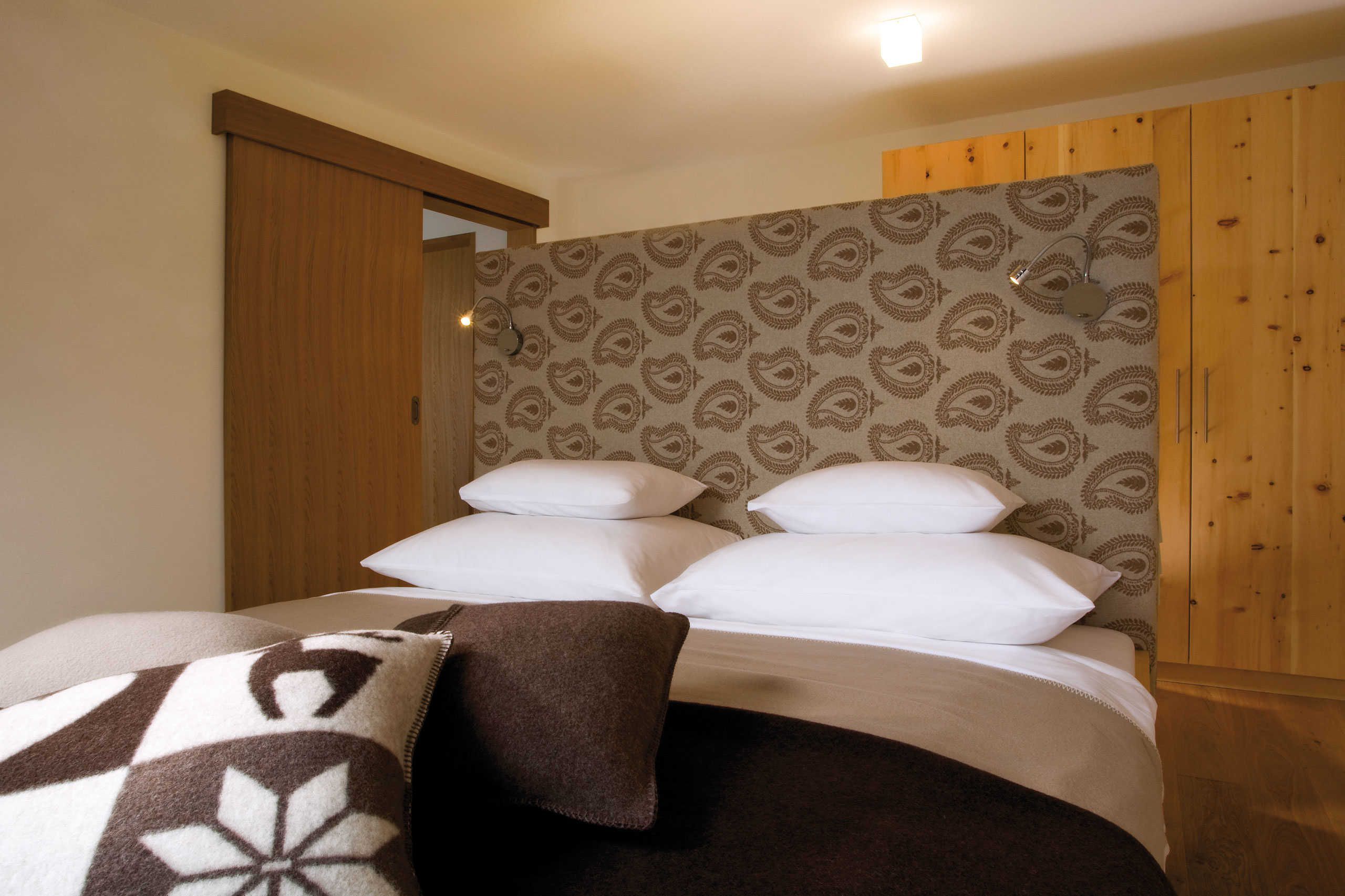 A bedroom with a bed, pillows, a blanket, wooden walls, and a patterned wallpaper.
