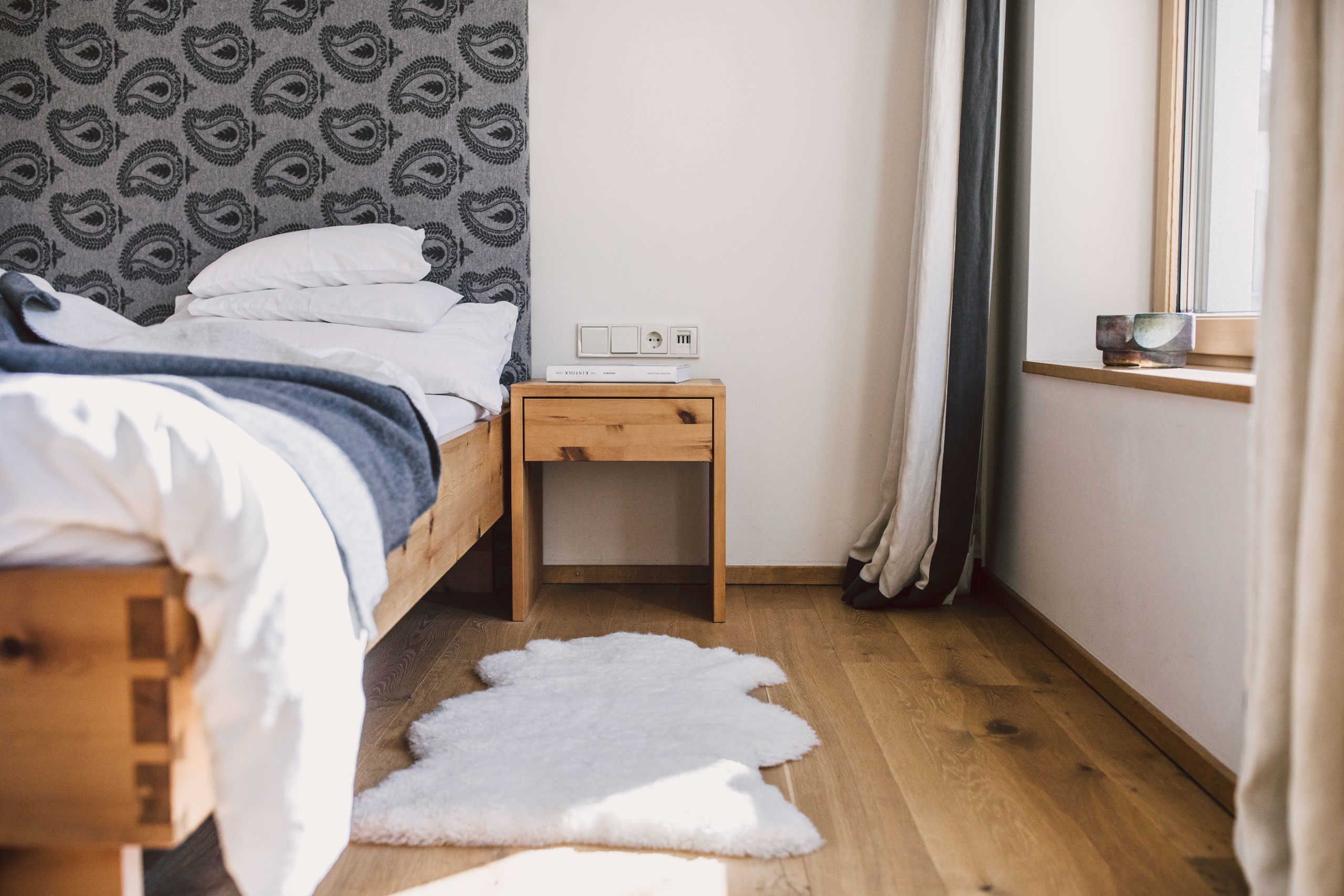 A bedroom with a wooden bed, a bedside table with a power outlet, a white rug, and a window with curtains.