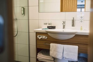 Ein Badezimmer mit weißem Waschbecken, Holzschrank, Spiegel, Handtuchhalter und einer Duschkabine aus Glas.