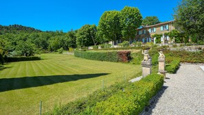 Un jardin bien entretenu, avec une pelouse, des haies et un bâtiment doté d’un toit en tuiles et de volets bleus.