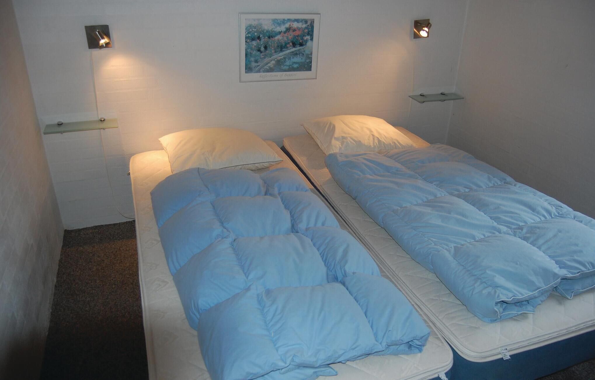 En säng för två personer med blått sänglinne i ett rum med vita väggar, en tavla på väggen och två vägglampor.