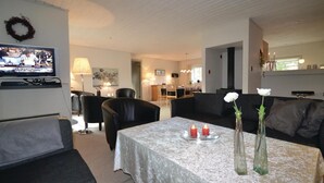 Una sala de estar con una televisión, dos sofás de cuero negro, una mesa de centro con mantel blanco y dos jarrones de vidrio con flores blancas.