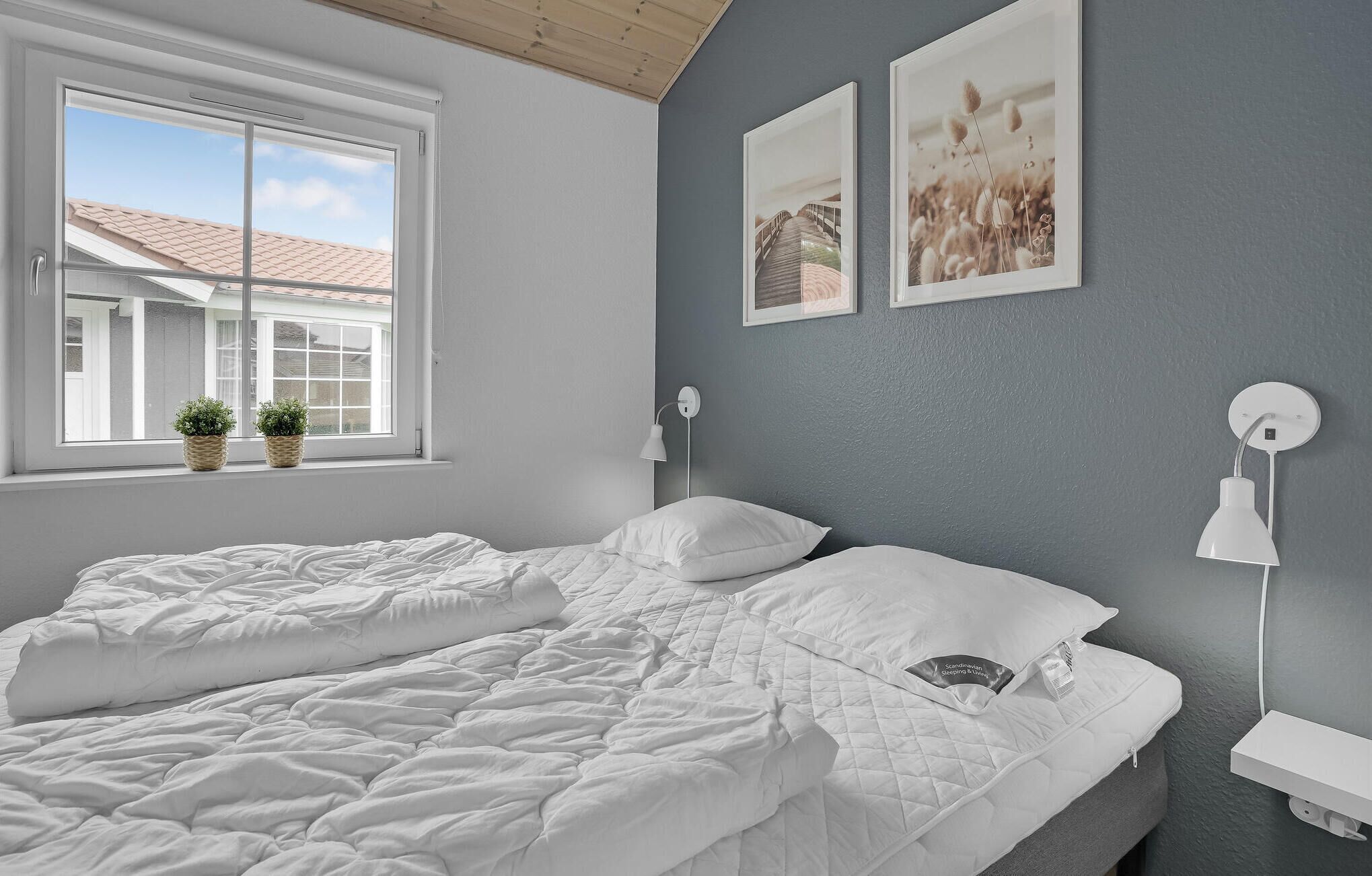 A bedroom with a large bed, two bedside tables, a window with plants, and two wall-mounted lamps.