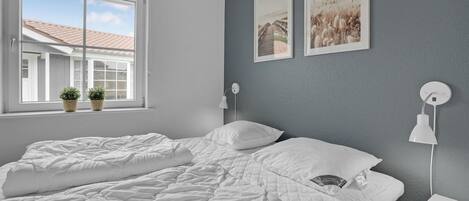A bedroom with a large bed, two bedside tables, a window with plants, and two wall-mounted lamps.