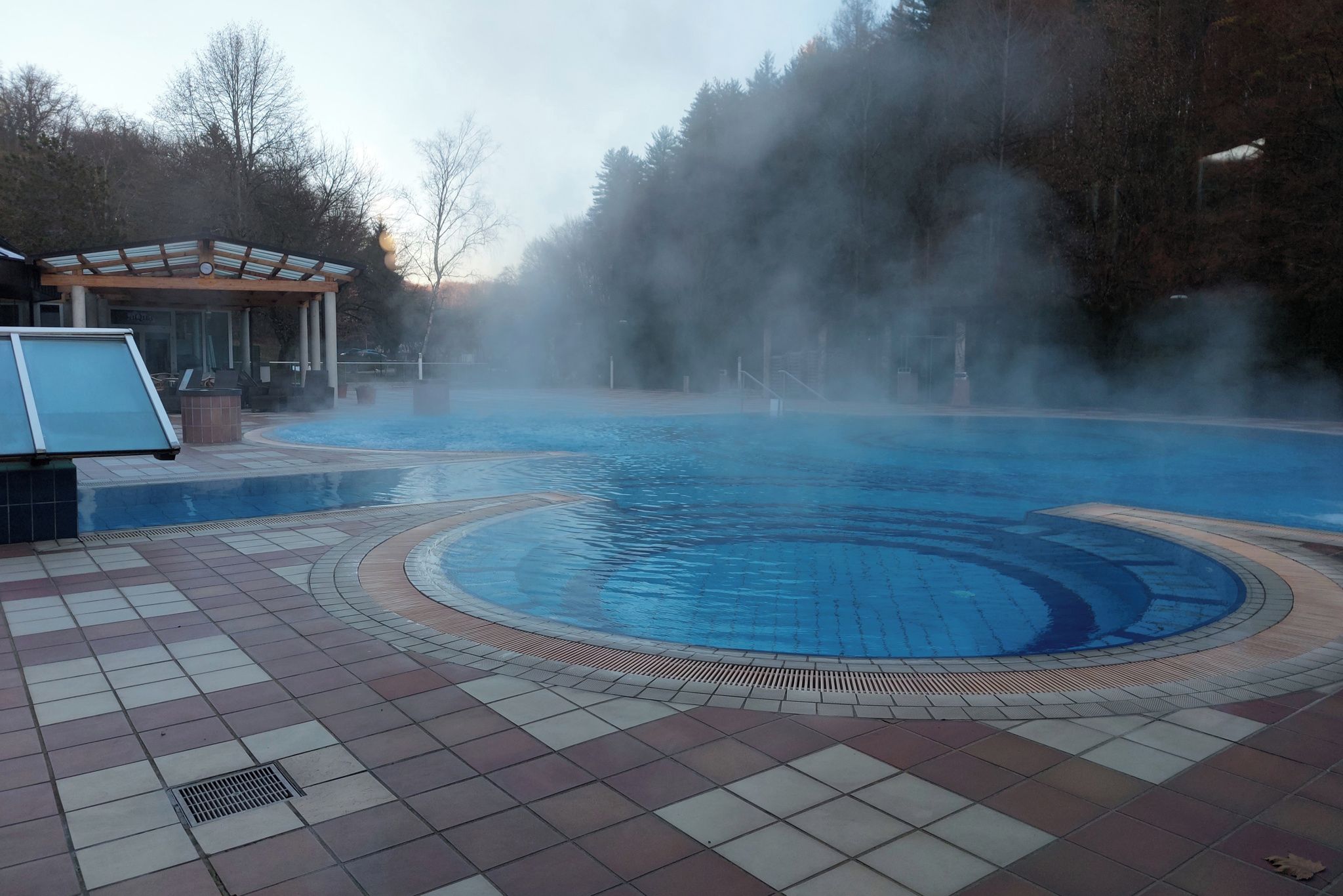 A swimming pool with a tiled deck, a covered area with a bench, and steam rising from the water.