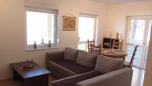 Uma sala de estar com sofá, mesa pequena e mesa de jantar com cadeiras. São visíveis janelas e uma porta.