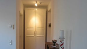 Um corredor estreito com piso de madeira, uma porta fechada no final e uma pequena mesa com um objeto decorativo à direita.