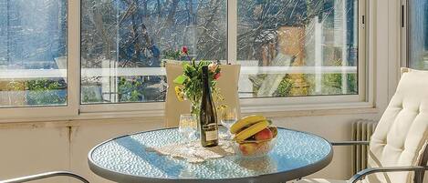A round glass table with a bottle of wine, a vase with flowers, and a bowl of fruit.
