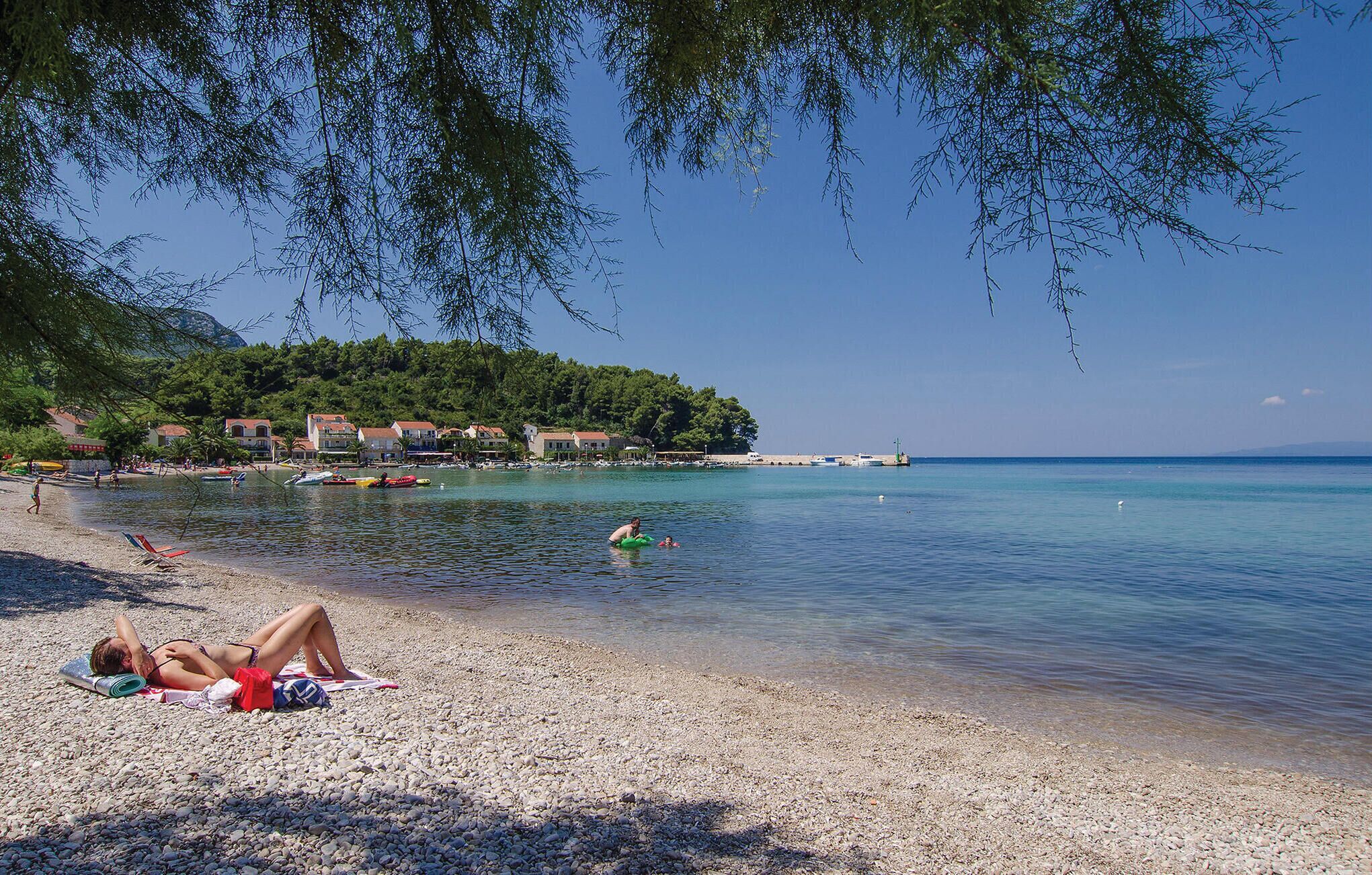 Une plage de galets avec une eau claire, une personne qui prend un bain de soleil et des bâtiments en arrière-plan.