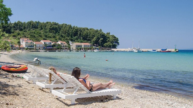 A beach with white lounge chairs, a clear blue sea, and buildings in the background. - Lovely apartment in Zuljana with house sea view (Zuljana)