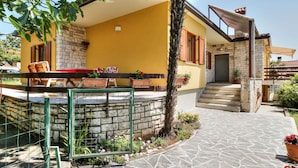 Un edificio amarillo con fachada de piedra, un porche cubierto con mobiliario exterior y un camino de piedra que conduce a una escalera.