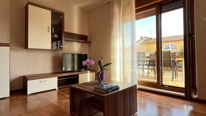 Amplia sala de estar con piso de madera, una mesa de comedor con un florero, un televisor y un gran ventanal corredizo que da a un patio exterior.