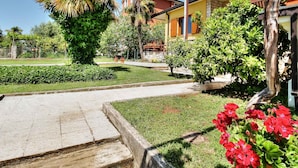 Una zona residencial con un jardín cuidado, un sendero que lleva a un edificio con balcón, y un cielo despejado.