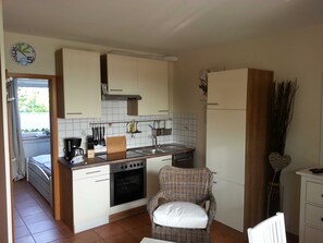 Eine kompakte Küche mit weißen Schränken, einem Backofen, einer Spüle und einem Essbereich mit einem Korbstuhl.