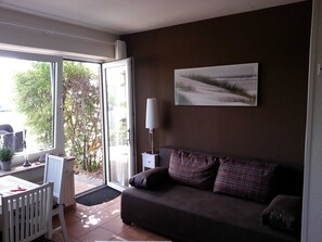 Un salon avec un canapé, une table et une chaise. Il y a une fenêtre donnant sur des arbres et on peut voir une voiture à l’extérieur.