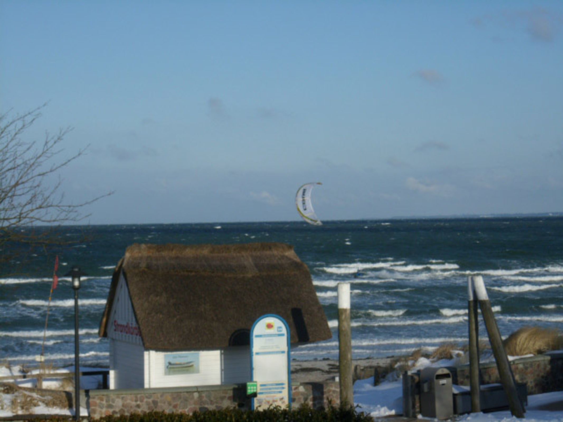 A coastal building with a thatched roof, a signboard, and a view of the sea with a kite surfer in the distance.