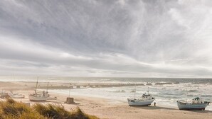 Een strand met meerdere boten afgemeerd in de buurt van de kust, een steiger die het water in reikt, en een bewolkte lucht.