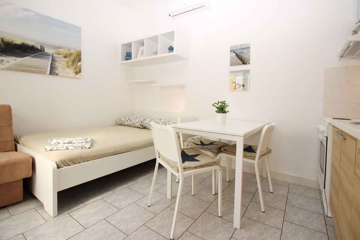 A compact room with a bed, a small dining table, and chairs. There are shelves with decorative items and a potted plant.