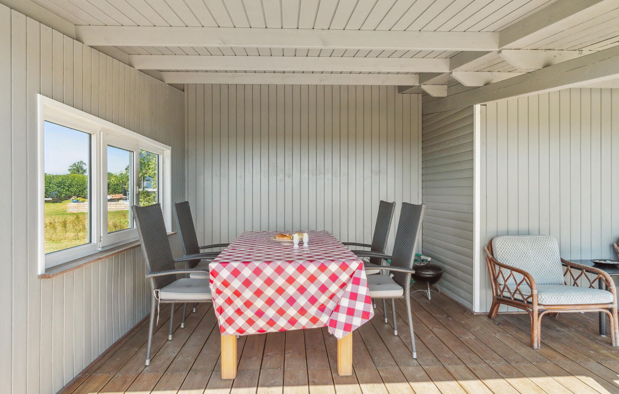 Un espace extérieur abrité, aménagé pour les repas, avec une table dressée pour quatre personnes, entourée de chaises, et une chaise en osier placée sur le côté.