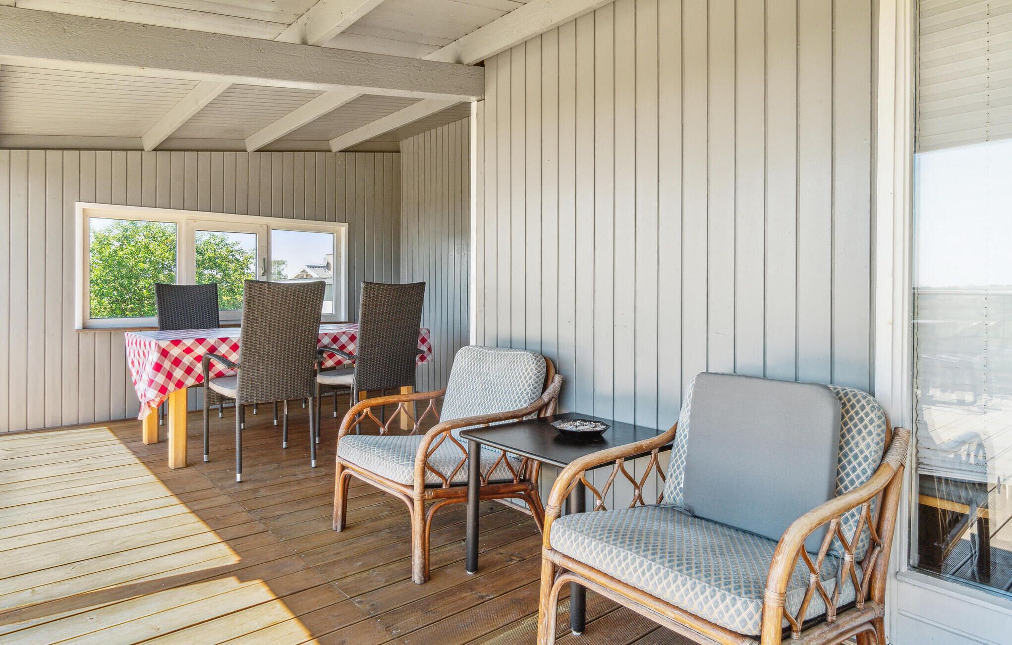 A covered outdoor seating area with wicker chairs and a table, wooden flooring, and a view through the window.