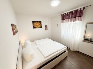 A bedroom with a bed, a window with curtains, a wall-mounted mirror, and artwork.