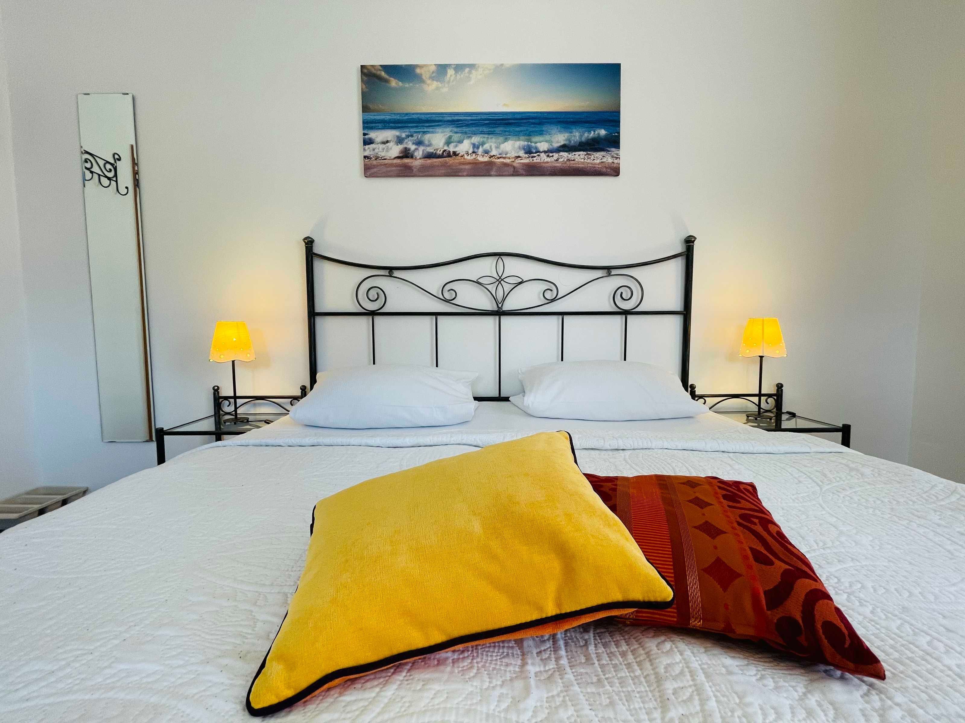 A neatly made bed with two pillows, a yellow pillow, and a red patterned pillow. There is a painting of a beach scene above the bed.