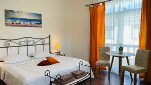 Quarto de hotel com cama, mesa de cabeceira com luminária, cadeira, pequena mesa com planta e janela com cortinas.