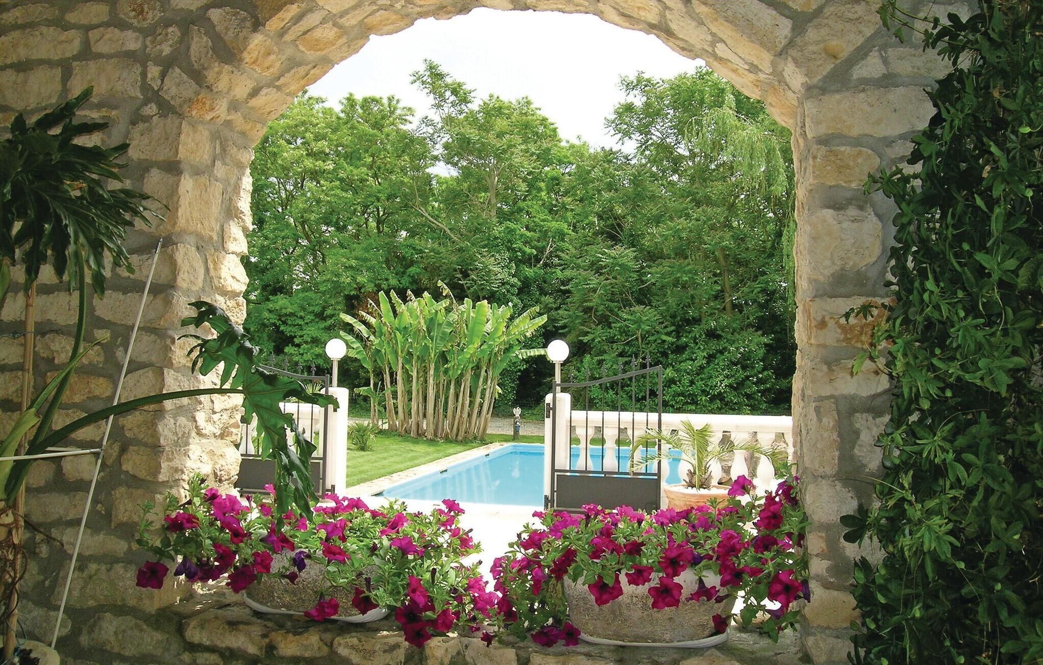 An outdoor pool area with a stone archway, surrounded by lush greenery and flowering plants.