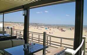 Vista para a praia a partir de uma área coberta, com mesas e cadeiras, com vista para uma praia cheia de guarda-sóis e pessoas.