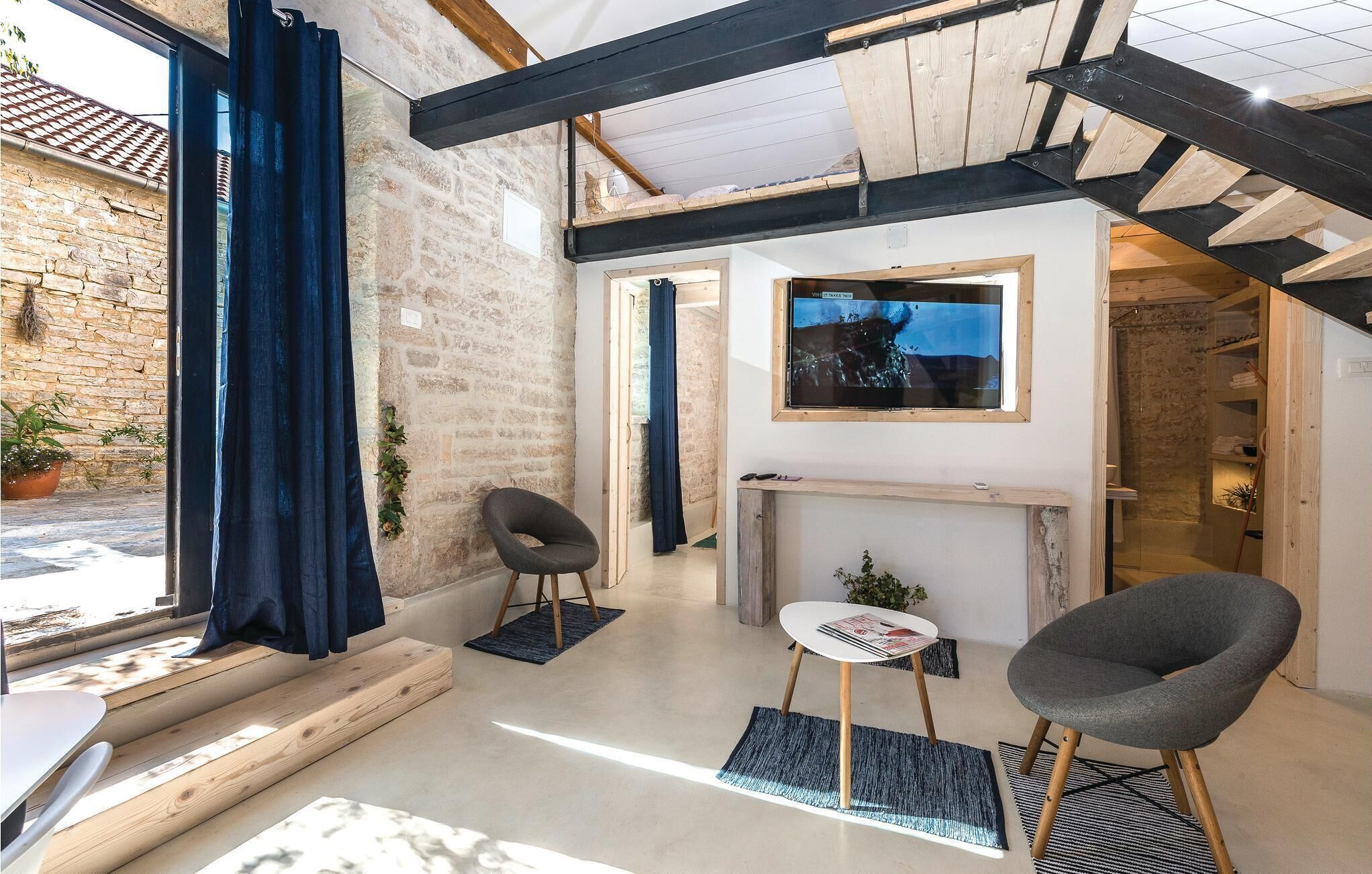 A modern living room with exposed beams, a stone wall, a fireplace, and a TV mounted on the wall.