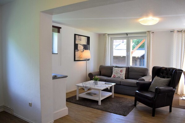 A living room with a sofa, armchair, coffee table, lamp, and a window with curtains. - Vacation apartment Timmi 4, 2-room apartment for 4 persons, ground floor - Ocean Way House (Scharbeutz)