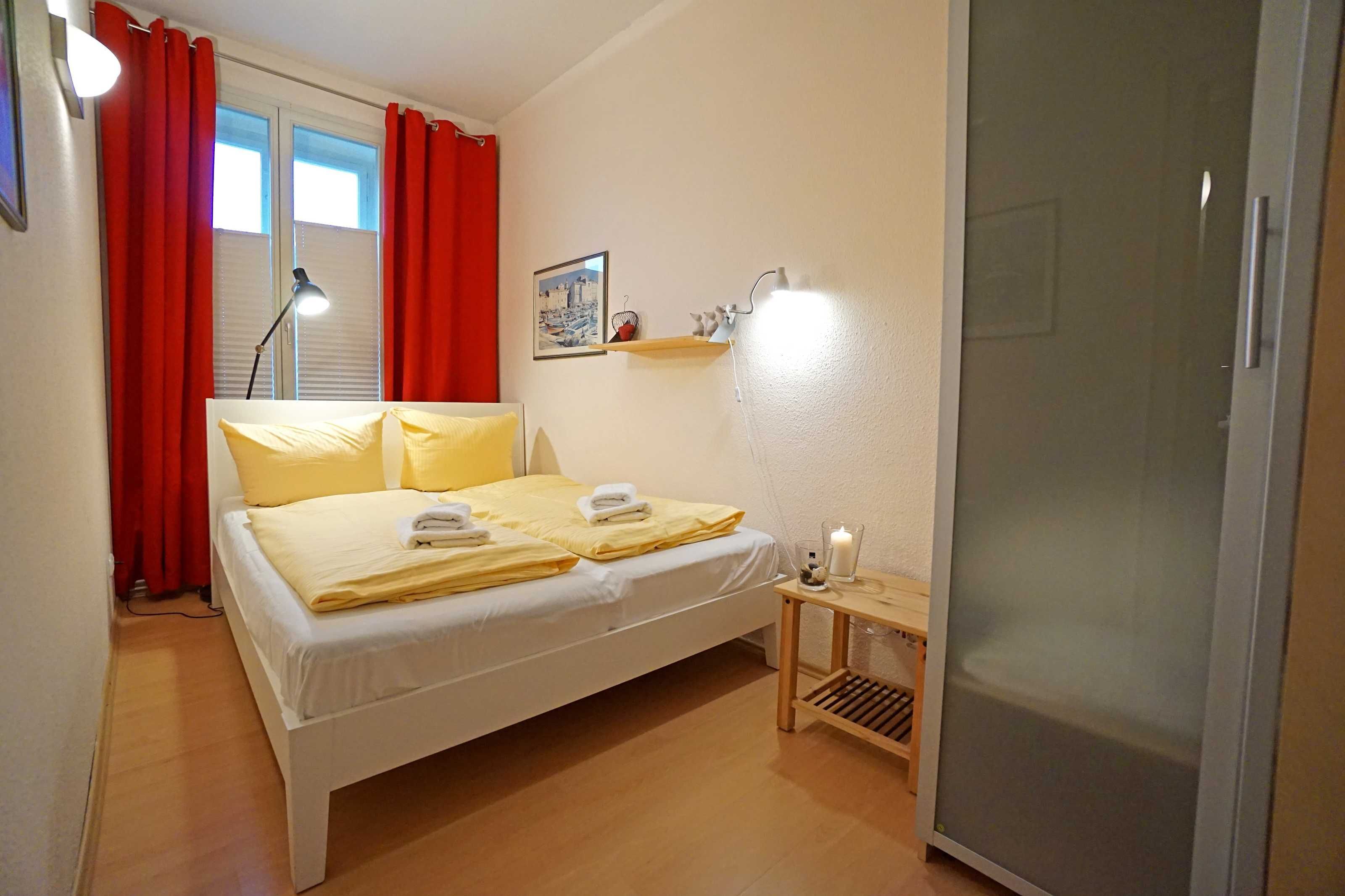 Ein ordentlich bezogenes Bett mit gelber Bettwäsche, ein Nachttisch mit Lampe und ein Fenster mit roten Vorhängen.
