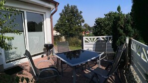 A patio area with a table and chairs, a barbecue grill, and a white fence. - Vacation cottage Robert, Germany (Bergen auf Rügen)