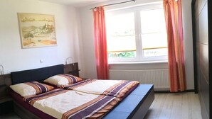 A bedroom with a bed, a window with curtains, a radiator, and a painting on the wall. - Vacation cottage Robert, Germany (Bergen auf Rügen)