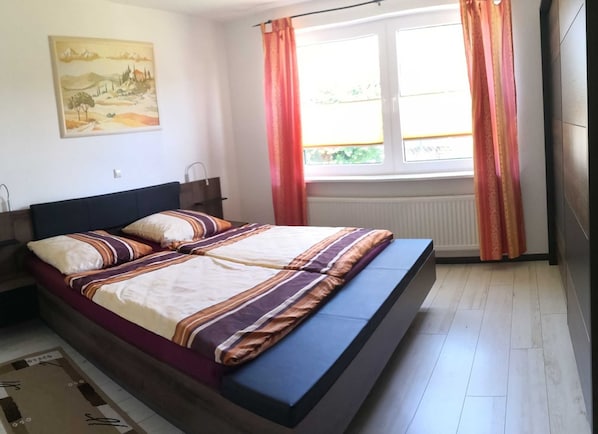 A bedroom with a bed, a window with curtains, a radiator, and a painting on the wall. - Vacation cottage Robert, Germany (Bergen auf Rügen)