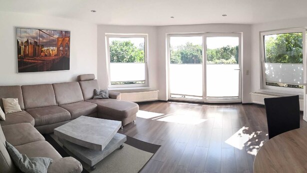 A modern living room with a sectional sofa, a coffee table, a round dining table, and a TV. There are large windows providing natural light and views of greenery. - Vacation cottage Robert, Germany (Bergen auf Rügen)