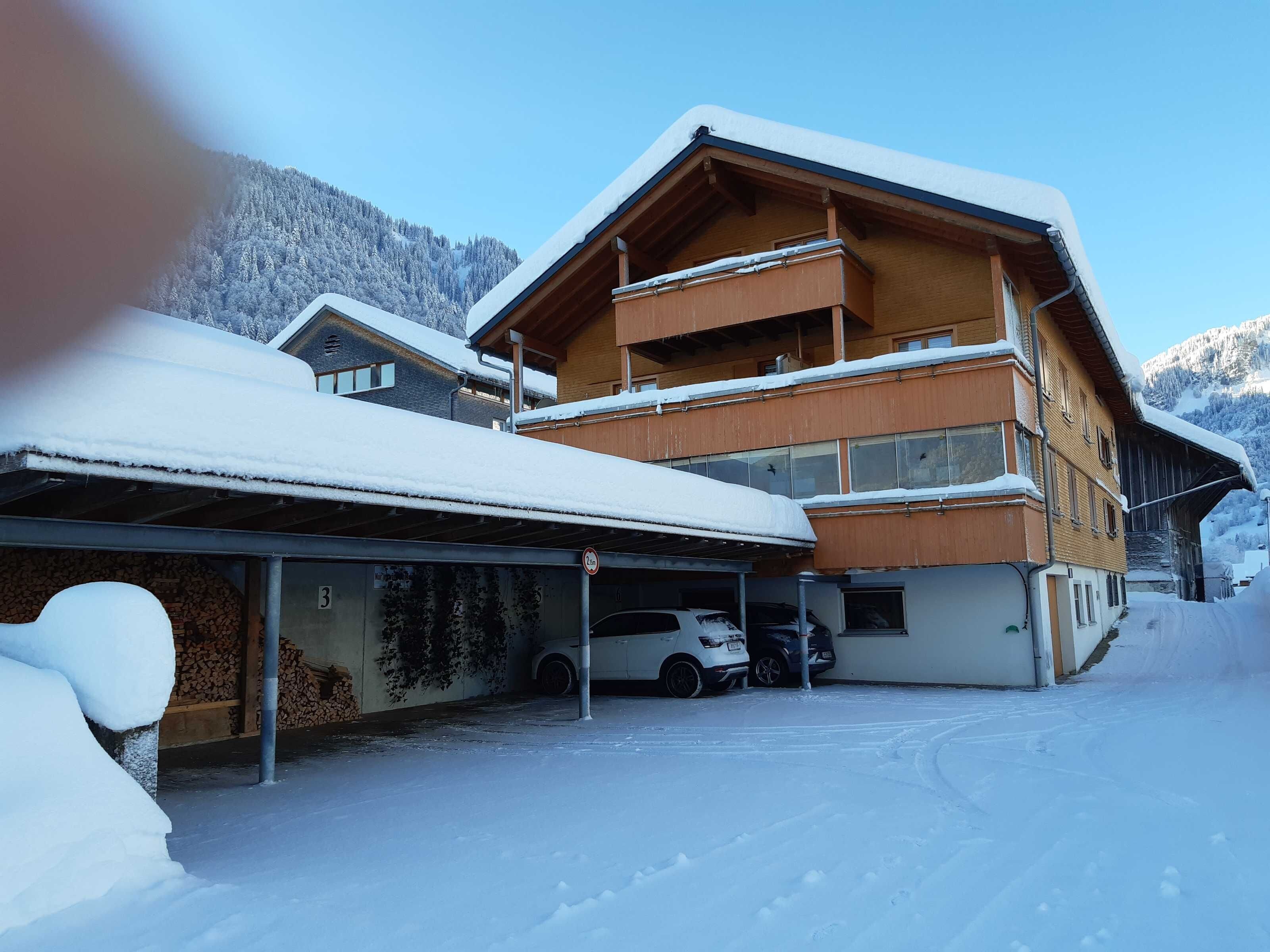 Un edificio de varios pisos con fachada de madera, zona de estacionamiento cubierta y montañas nevadas al fondo.