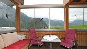 Ein Zimmer mit Holzbalkendecke, einem rot gestreiften Sofa, zwei roten Sesseln, einem kleinen Tisch und großen Fenstern mit Blick auf die Berge.