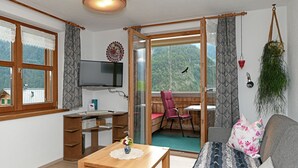 A room with a wooden desk, a TV, a sofa, and a sliding glass door leading to a balcony with a chair.