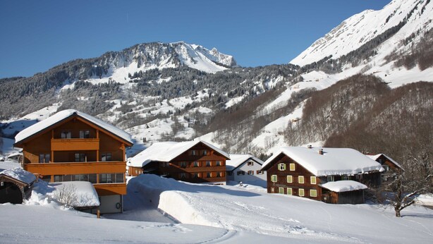 A snow-covered mountain village with wooden buildings and a prominent mountain in the background.
