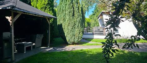 A covered outdoor seating area with a table and chairs, surrounded by a well-maintained lawn and tall trees.