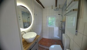 A bathroom with a white sink, a mirror with a circular light, a shower, and a window.